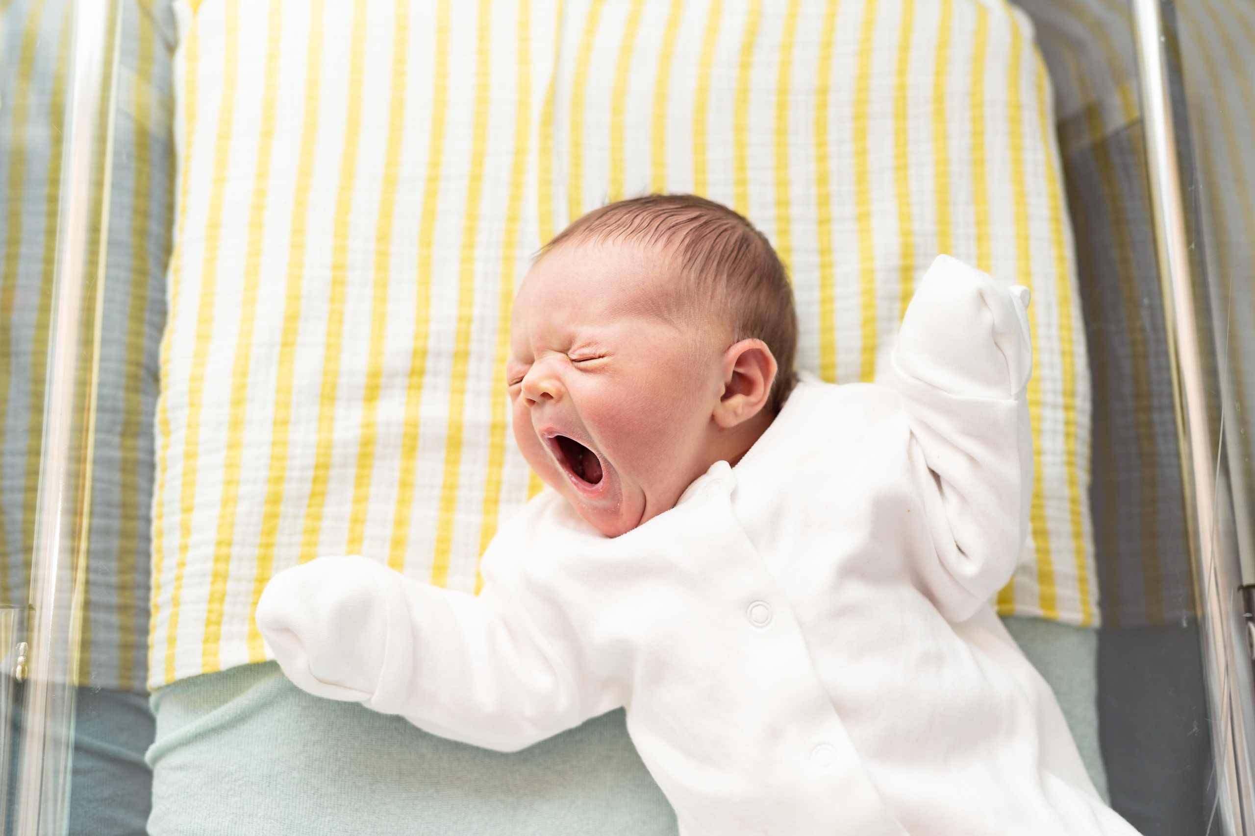 Newborn baby yawning, lying in a hospital crib.